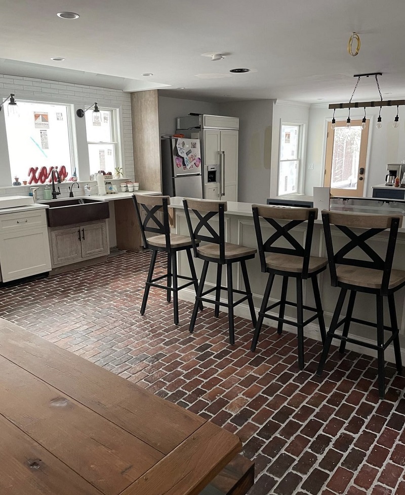 Rustic kitchen and dining area with a red brick floor, white cabinetry, a wooden farmhouse table, and black bar stools