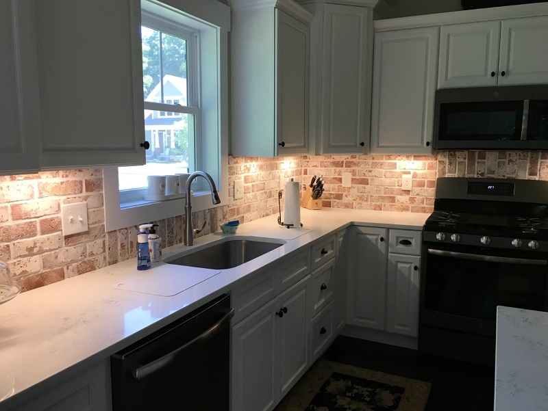 U-shaped kitchen with white cabinetry and countertops, featuring rustic brick backsplashes and under-cabinet lighting